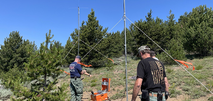 Caucasian men in a field between green trees setting up large metal poles with tripod-like support